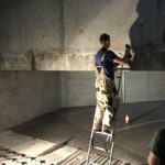 Mężczyzna na drabinie wykonuje iniekcje krystaliczne na betonowej ścianie.