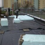 Robotnicy montują izolacje przeciwwilgociowe na placu budowy przed blokiem mieszkalnym.