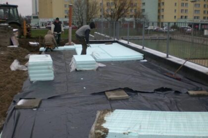 Robotnicy montują izolacje przeciwwilgociowe na placu budowy przed blokiem mieszkalnym.