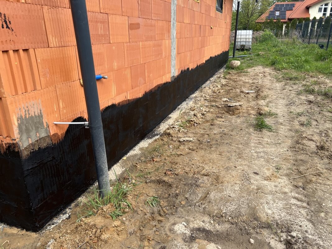 Ściana budynku z wyraźnie widoczną izolacją fundamentów w postaci ciemnej powłoki.