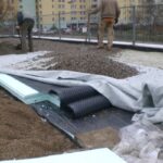 Robotnicy układają warstwy materiałów, w tym izolacje przeciwwilgociowe, na placu budowy.