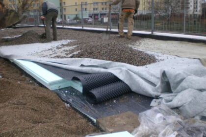 Robotnicy układają warstwy materiałów, w tym izolacje przeciwwilgociowe, na placu budowy.