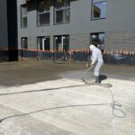 Pracownik w kombinezonie ochronnym wykonuje hydroizolację tarasu przy budynku.