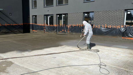 Pracownik w kombinezonie ochronnym wykonuje hydroizolację tarasu przy budynku.