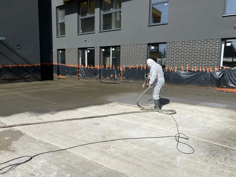Pracownik w kombinezonie ochronnym wykonuje hydroizolację tarasu przy budynku.