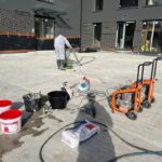 Pracownik wykonuje izolacje tarasów na dużej betonowej powierzchni przed budynkiem, korzystając z narzędzi i materiałów budowlanych.