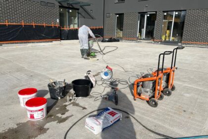 Pracownik wykonuje izolacje tarasów na dużej betonowej powierzchni przed budynkiem, korzystając z narzędzi i materiałów budowlanych.