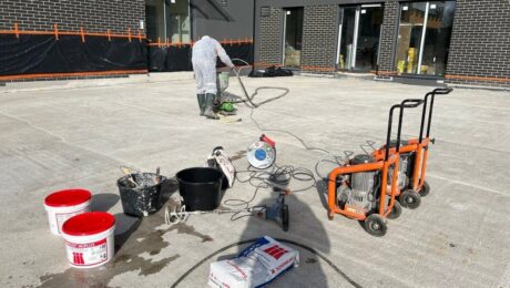 Pracownik wykonuje izolacje tarasów na dużej betonowej powierzchni przed budynkiem, korzystając z narzędzi i materiałów budowlanych.