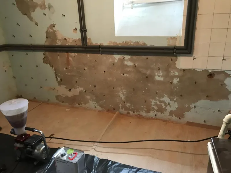 Ściana z pakerami i odbitym tynkiem pod oknem, na pierwszym planie pompa iniekcyjna