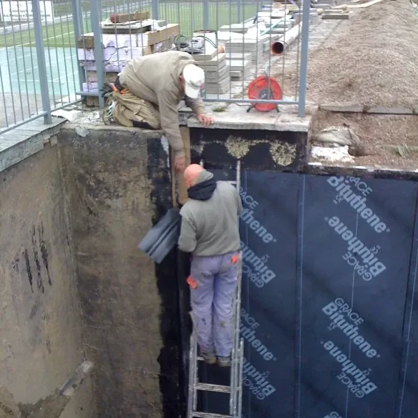 Pracownicy na drabinie przy klejeniu membrany na ścianę fundamentową w wykopie