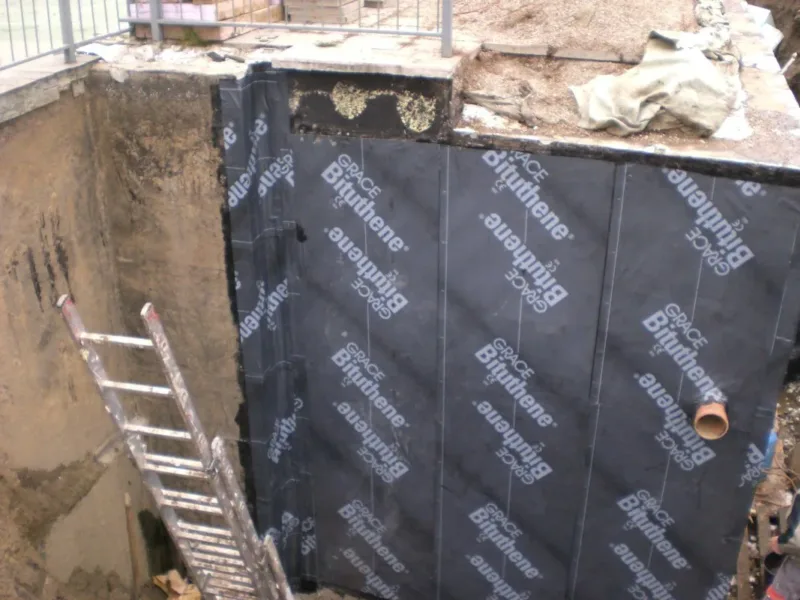 Odkopana ściana fundamentowa z nową membraną izolacyjną, w wykopie drabina