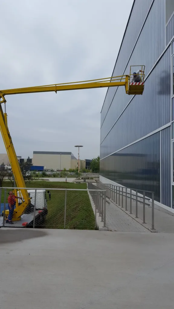 Podnośnik koszowy przy przeszklonej elewacji hali, na dole pochylnia z balustradą